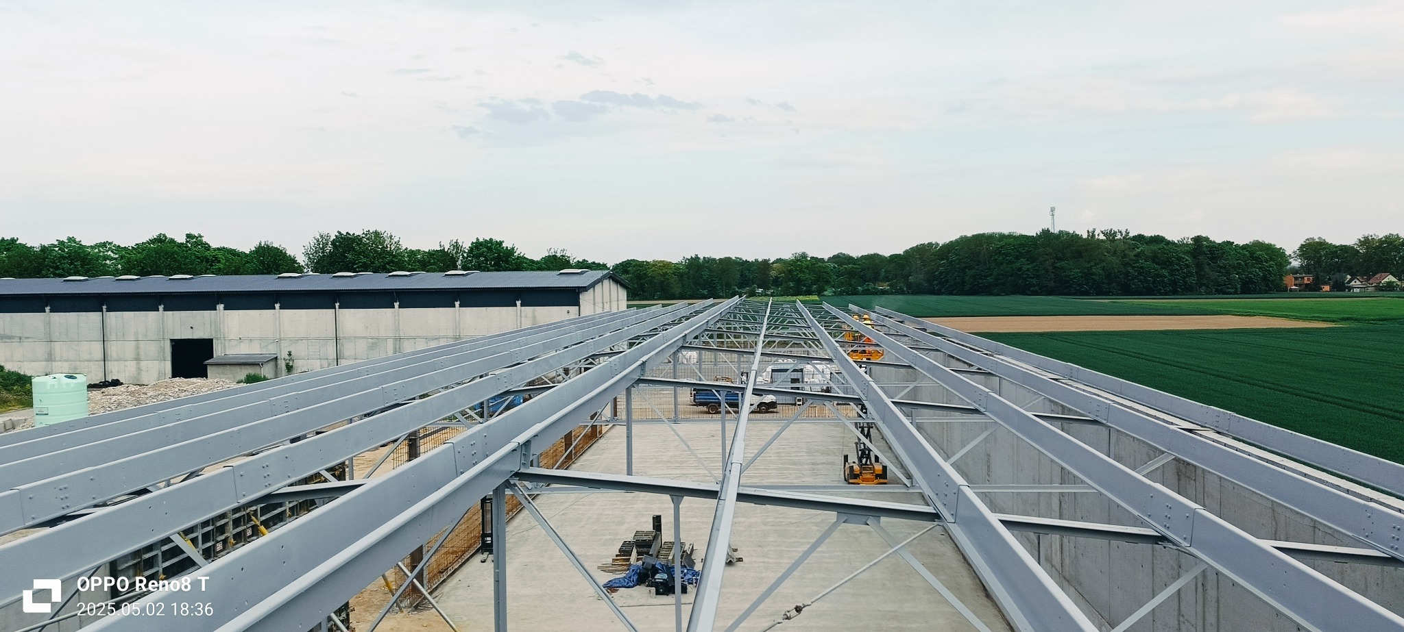 Konstrukcja stalowa hali w trakcie montażu, widok z góry. W tle budynek magazynowy i pola uprawne. Widoczne elementy montażowe i sprzęt budowlany.