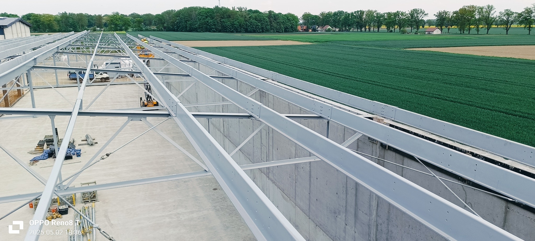 Konstrukcja stalowa hali w trakcie budowy, widok z góry na szkielet dachu. W tle zielone pola i las. Widoczne białe ściany i elementy konstrukcyjne.
