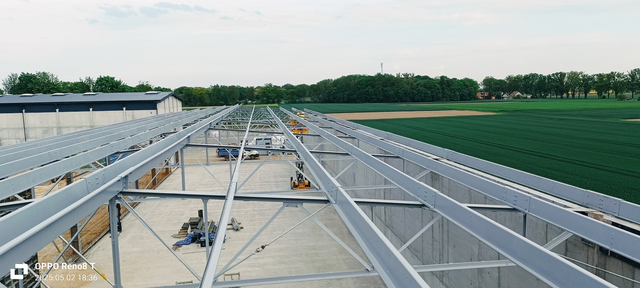 Konstrukcja stalowa hali w trakcie budowy, widok z góry na betonowe ściany, w tle zielone pola i las. Na placu budowy widoczne maszyny i materiały.