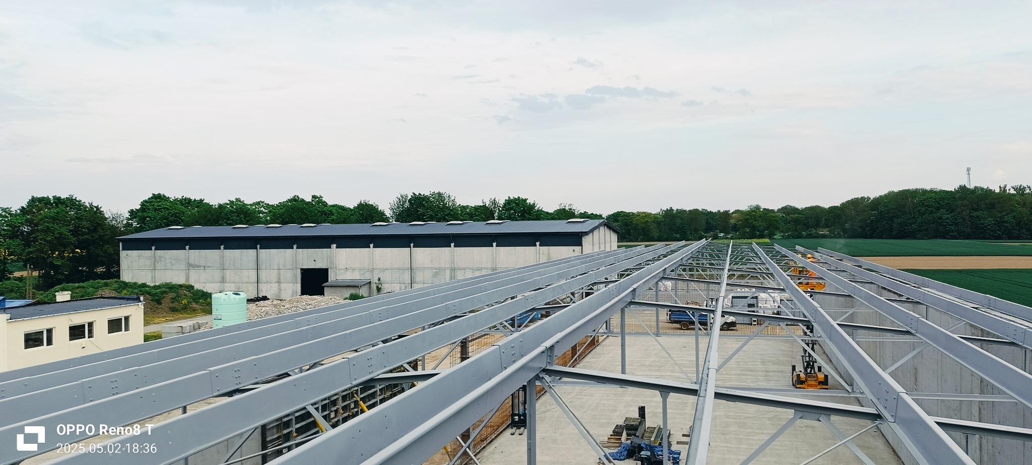 Konstrukcja stalowa hali produkcyjnej w trakcie montażu, widok z góry na szkielet budynku, betonowe ściany w tle, sprzęt budowlany na placu.