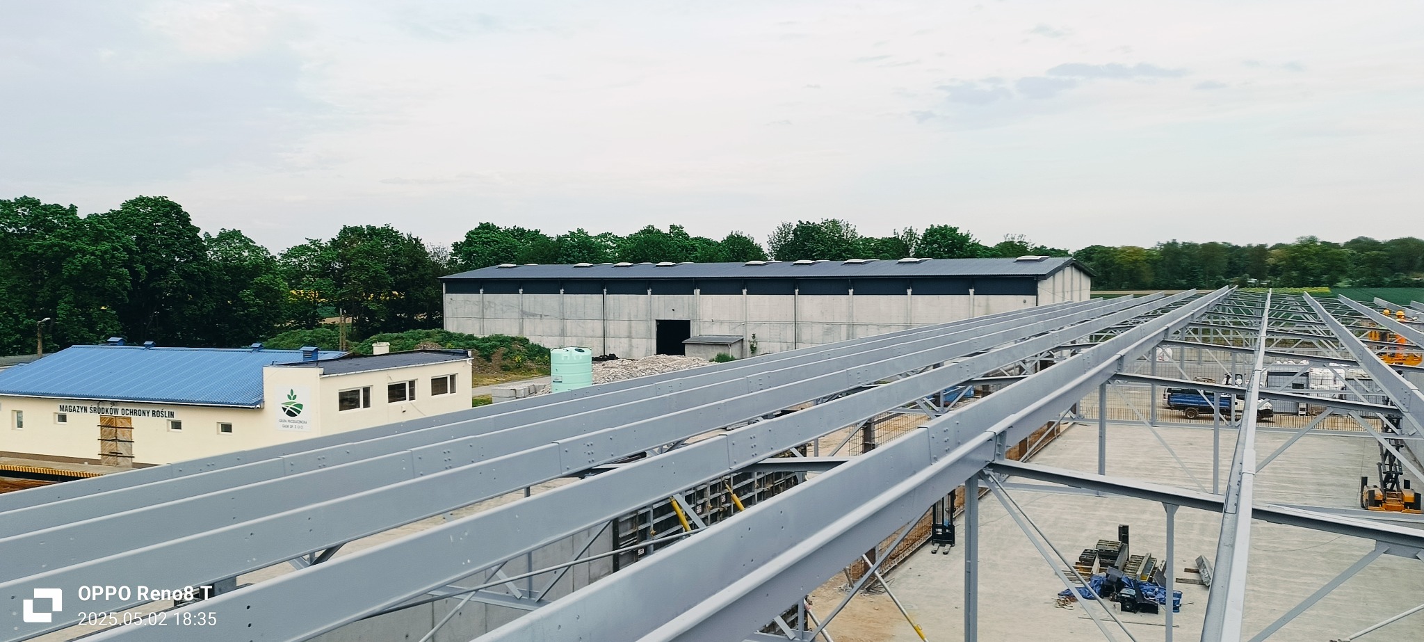 Konstrukcja stalowa hali w trakcie montażu, widok z góry. W tle budynek magazynowy i zieleń. Szara, pochmurna pogoda. Elementy konstrukcyjne pomalowane na szaro.