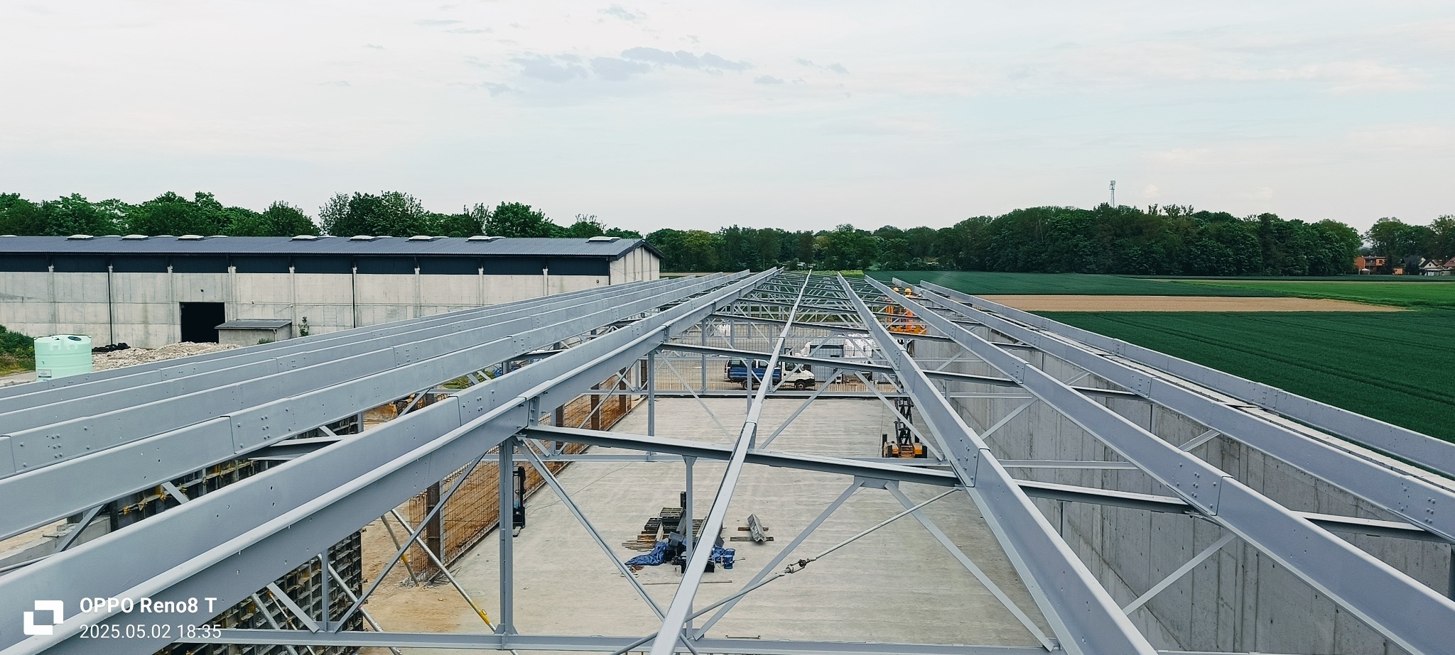 Wysoki kąt ujęcia szkieletu hali przemysłowej z betonu i stali na tle zielonych pól i budynków. Widoczna konstrukcja dachu i ściany. Prace w toku.