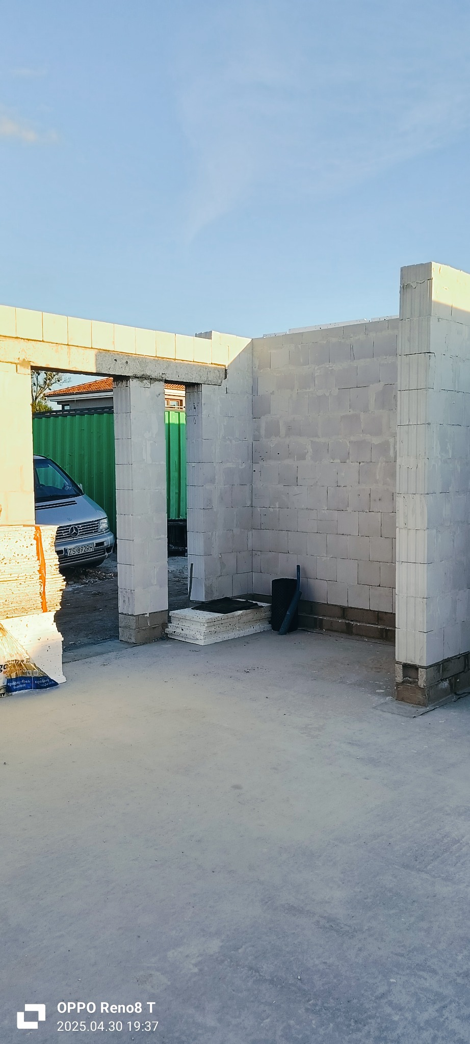 Wnętrze surowego budynku w trakcie budowy, z widocznymi ścianami z białych bloków i otworami drzwiowymi. Na zewnątrz zaparkowany srebrny samochód dostawczy.