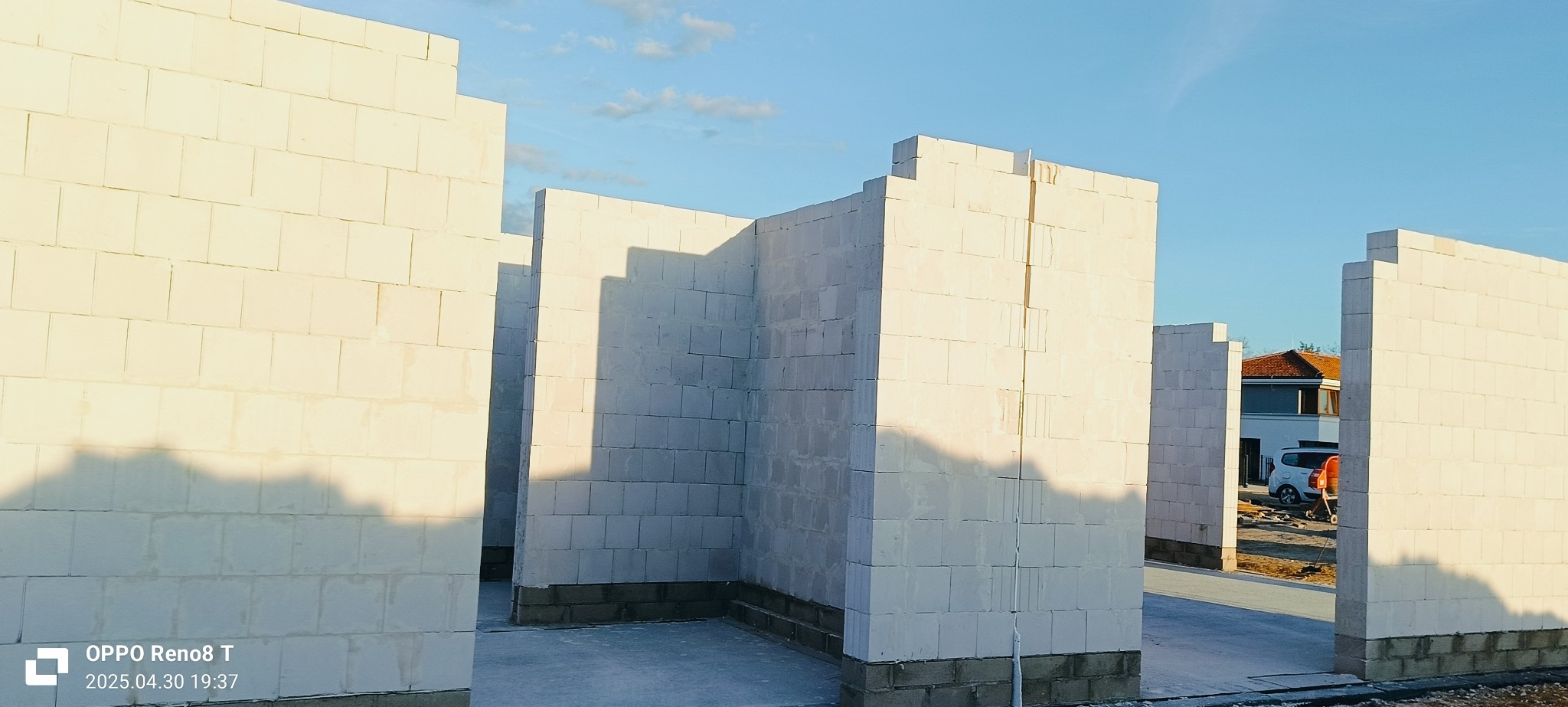 Wznoszone ściany z białych bloczków betonu komórkowego na fundamencie. Widoczne fragmenty sąsiedniego budynku i niebieskie niebo z delikatnymi chmurami.