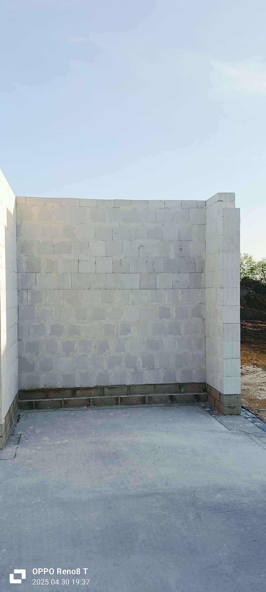 Wznoszone ściany z białych bloczków betonu komórkowego na fundamencie. Widoczny fragment terenu budowy i zieleń w tle. Kąt ujęcia z dołu podkreśla pionowość konstrukcji.