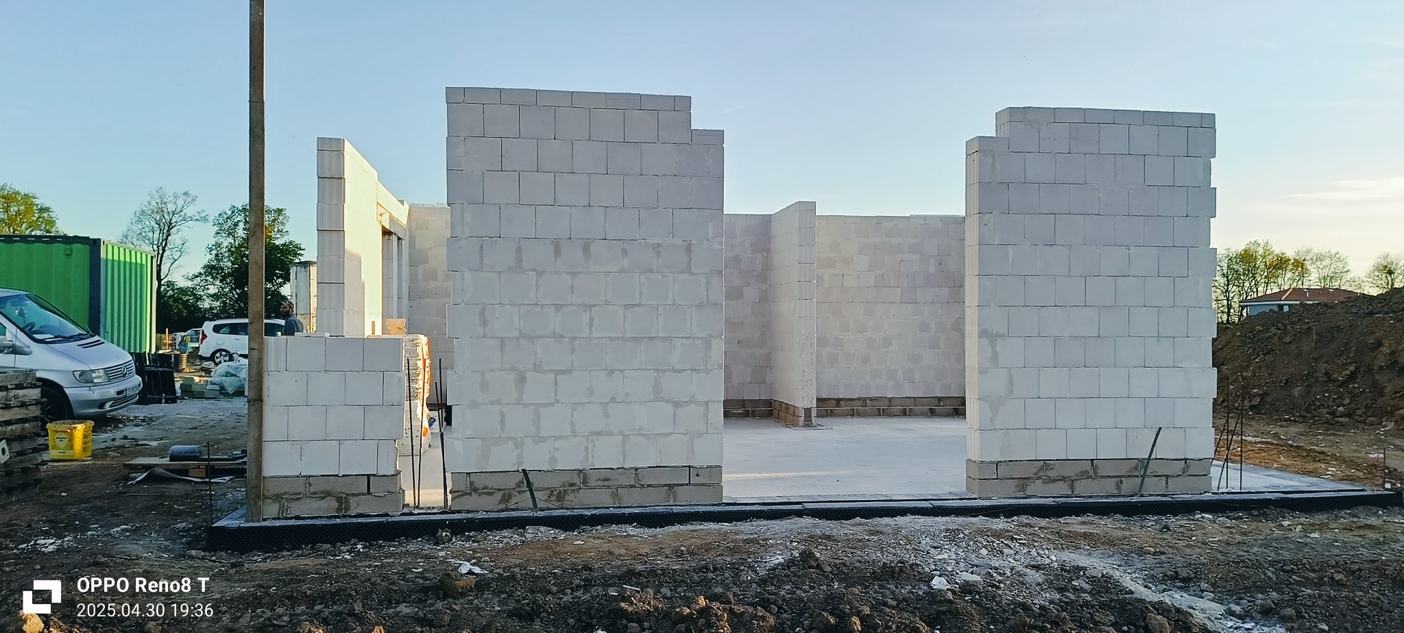 Wnętrze domu w trakcie budowy, ściany wzniesione z białych bloczków, widoczny fundament i teren budowy. W tle zaparkowane samochody i zieleń.
