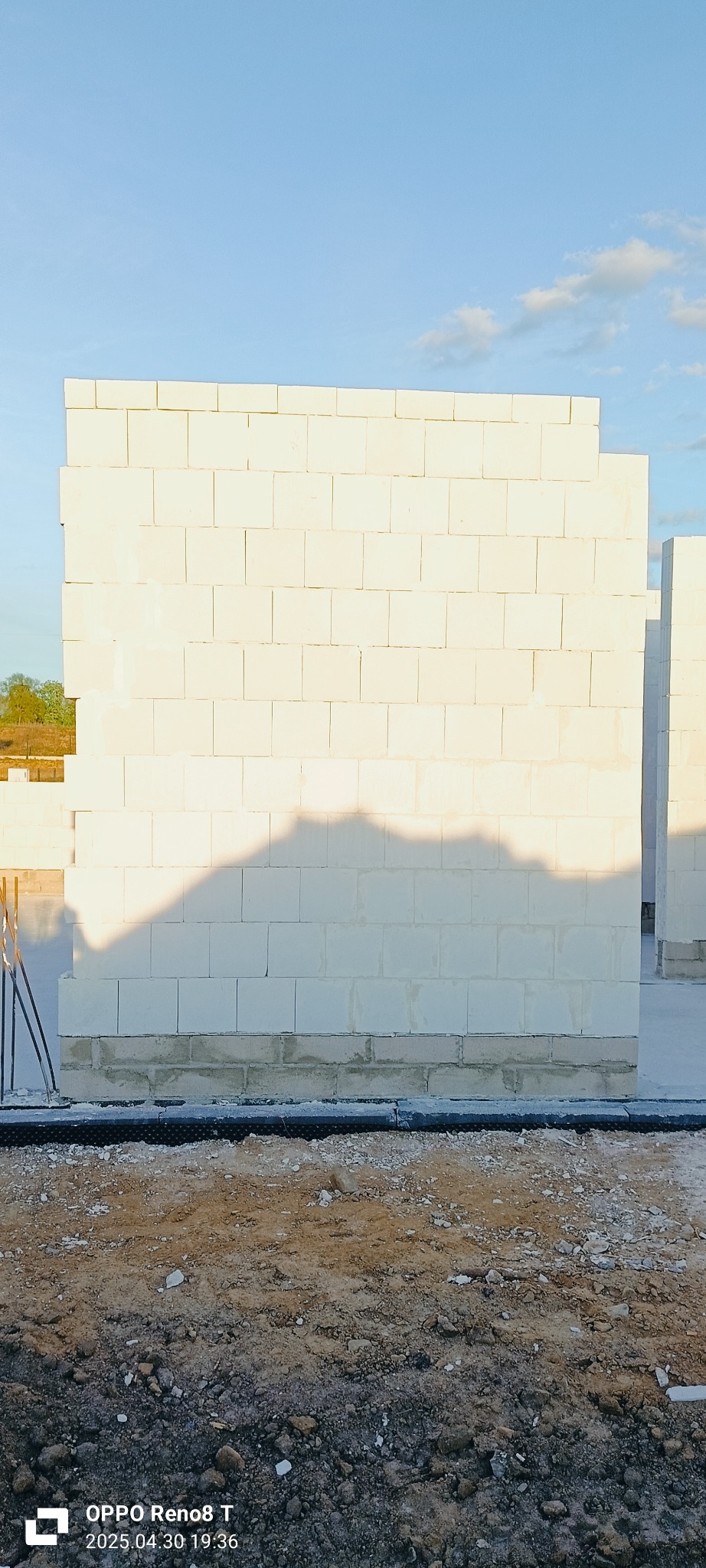 Wznoszenie ściany z białych bloczków na fundamencie. Widoczne zbrojenia i grunt. Ściana oświetlona słońcem, rzucająca cień. Częściowo widoczne niebieskie niebo.