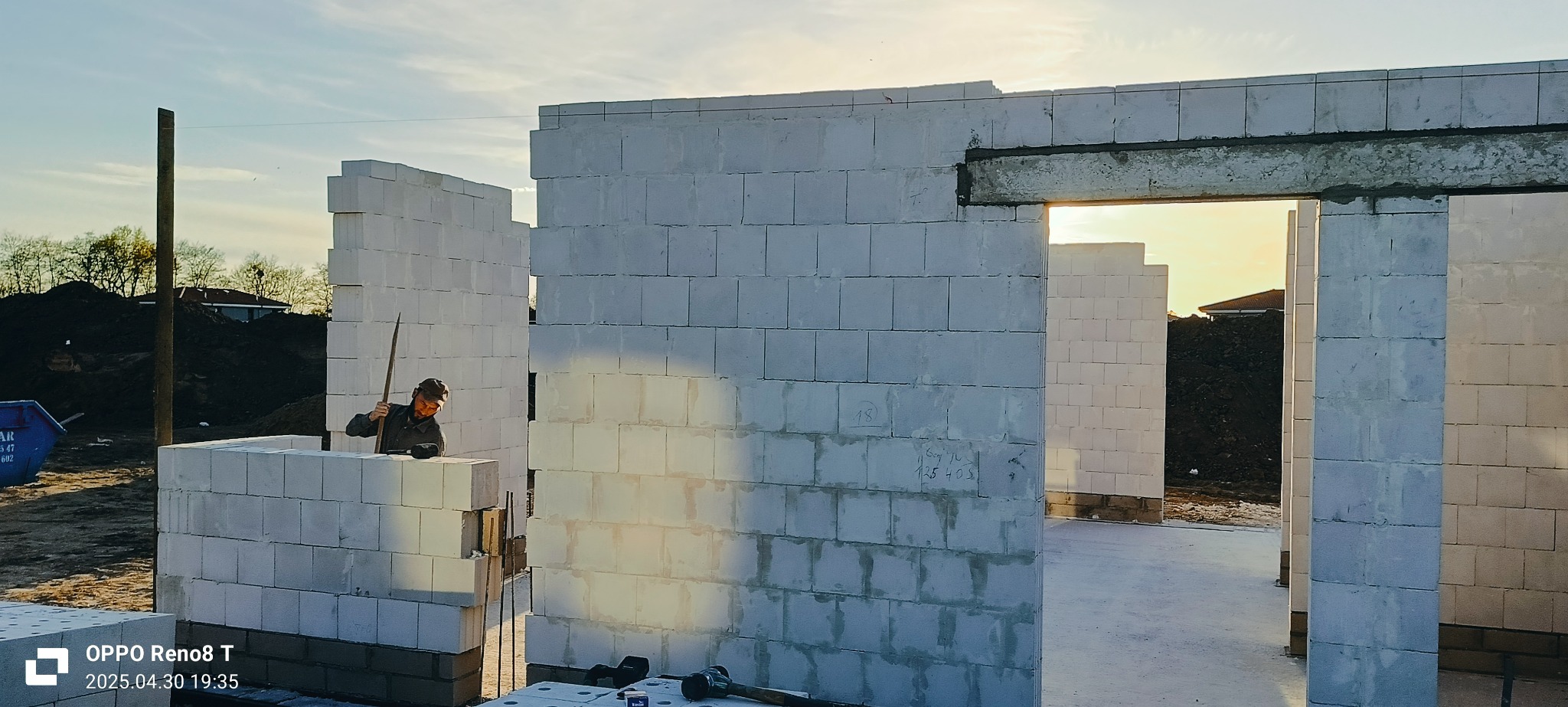 Wznoszenie ścian domu z białych bloczków, widoczny murarz przy pracy i częściowo wzniesiona konstrukcja w promieniach zachodzącego słońca.