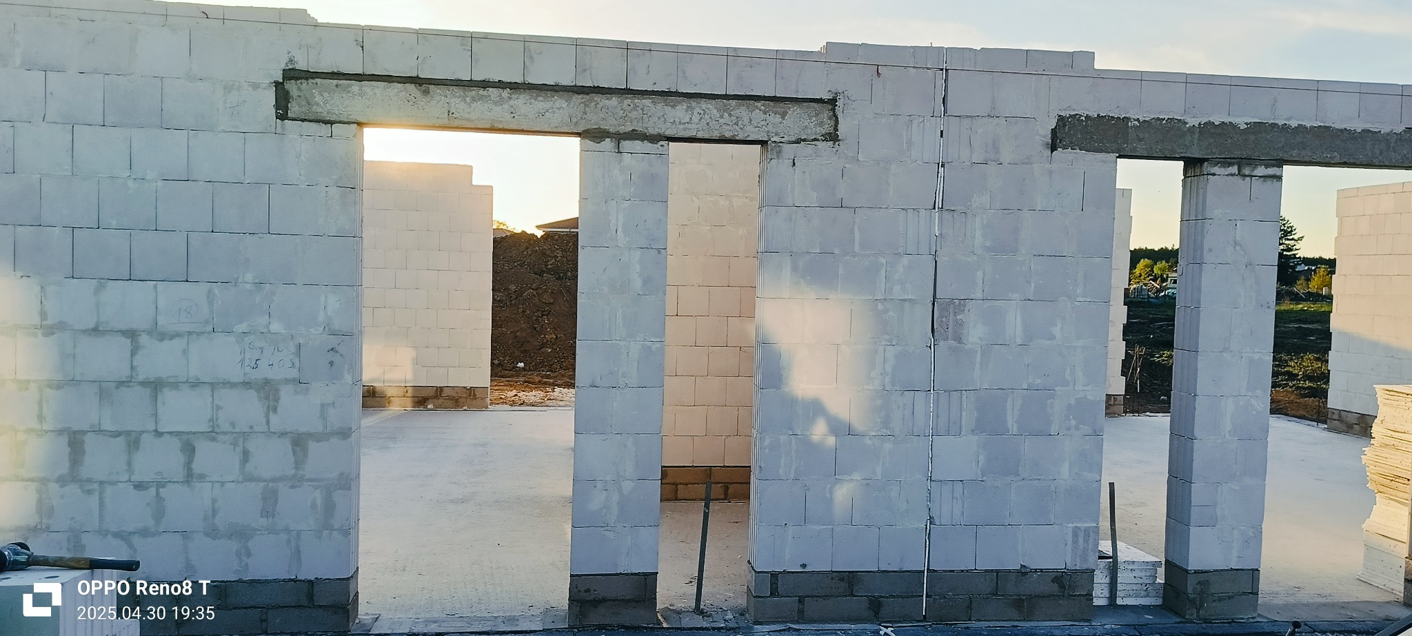 Wnętrze domu w trakcie budowy z białych bloczków, widoczne otwory okienne i drzwiowe z nadprożami, w tle ziemia. Etap murowania ścian z betonu komórkowego.