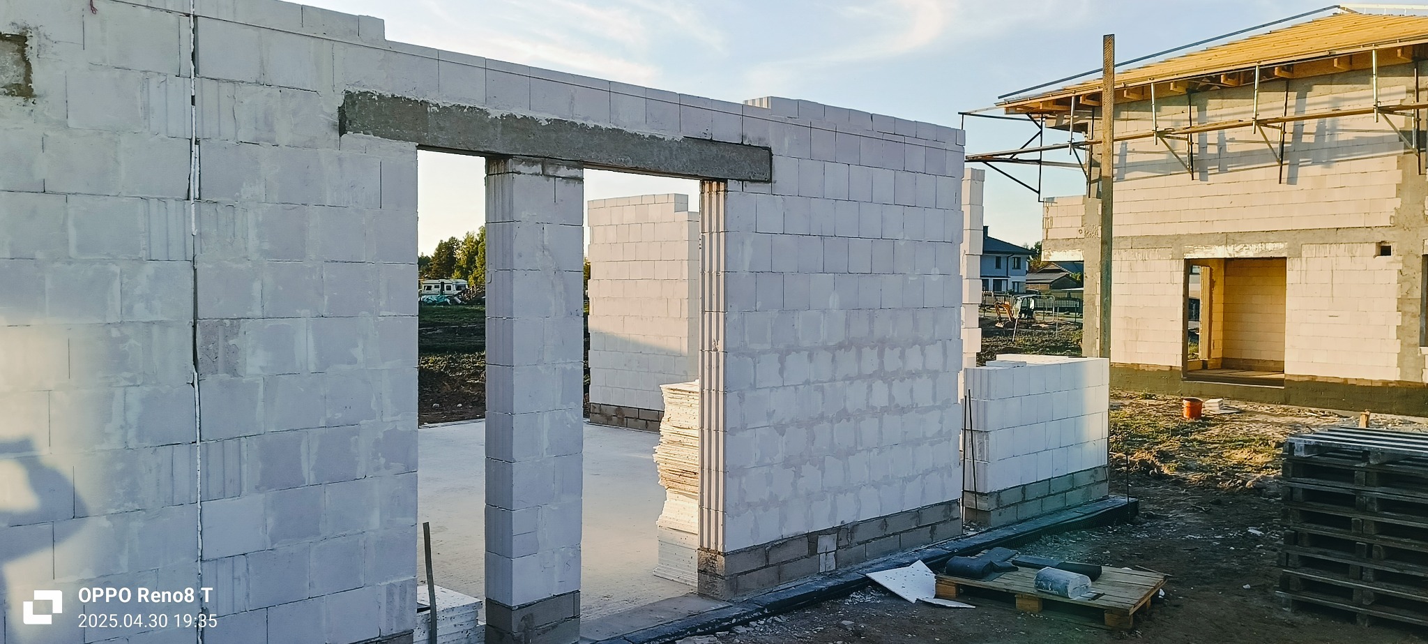 Wznoszenie ścian domu z betonu komórkowego. Widoczne nadproże nad otworem drzwiowym i paleta z bloczkami. W tle drugi budynek w trakcie budowy i zieleń.