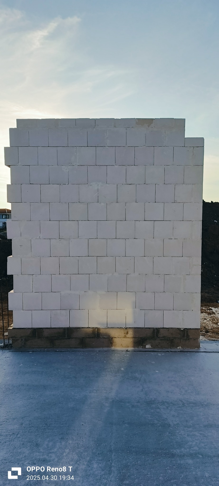 Wzniesiona ściana z białych bloczków na fundamencie, widoczna w świetle słonecznym. Fragment widocznego terenu i nieba w tle. Budowa domu w trakcie realizacji.