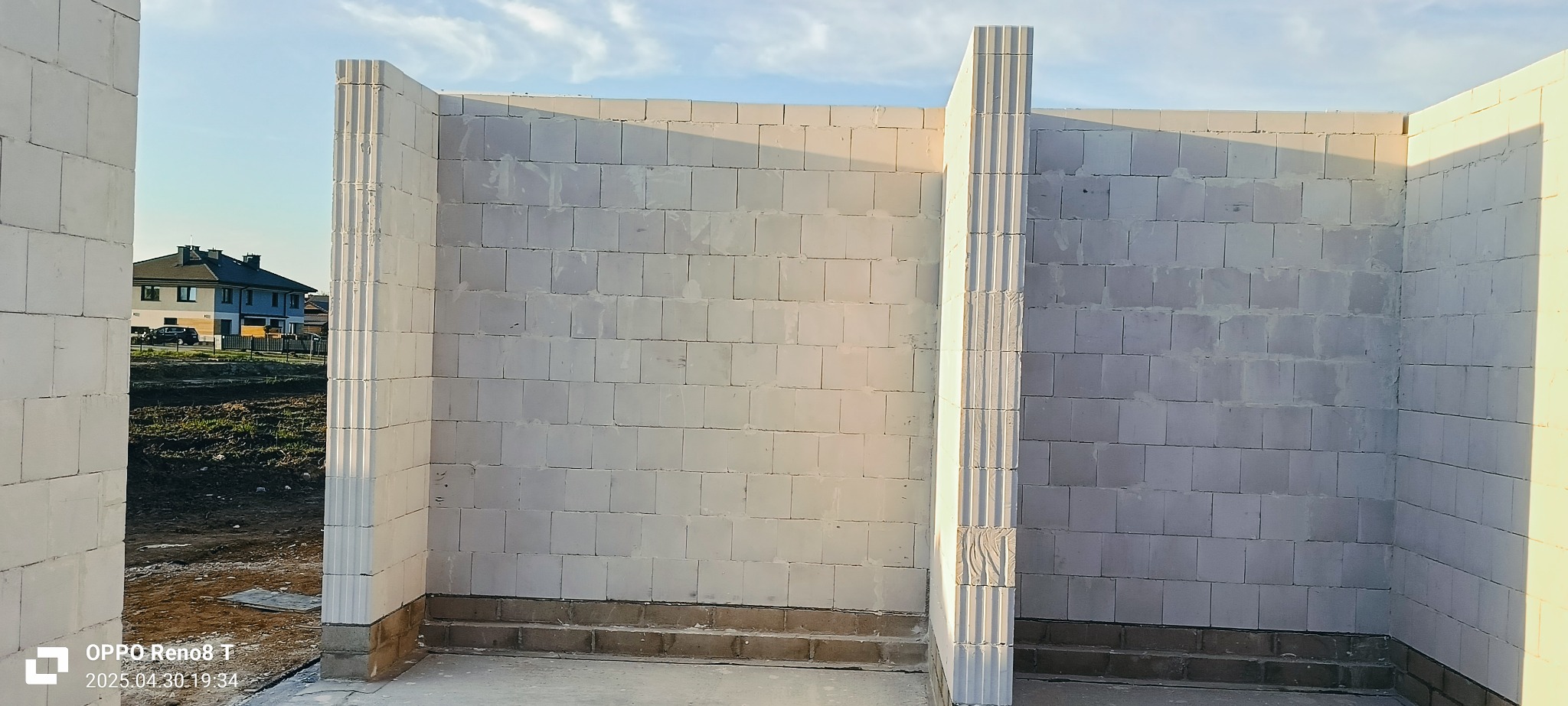 Wnętrze budynku w trakcie budowy z białych bloczków, widoczne ściany i fragment budynku w tle. Charakterystyczne pionowe elementy konstrukcyjne z żłobieniami.