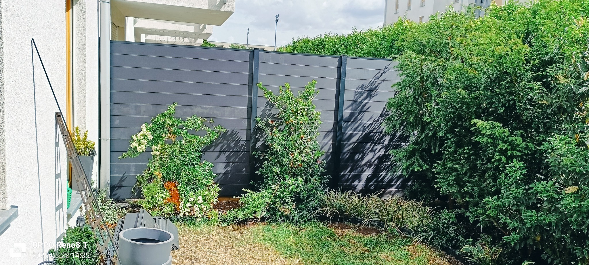 Nowoczesne ogrodzenie z paneli kompozytowych w kolorze grafitowym, częściowo przysłonięte przez bujną roślinność i kwiaty w ogrodzie.