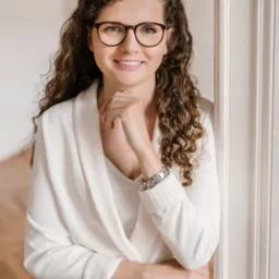 Portret uśmiechniętej kobiety w okularach i białym garniturze, opierającej się o białą ścianę z panelami.