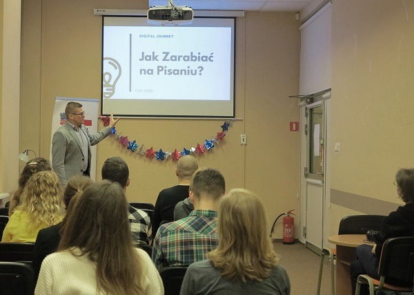 Prelegent w szarym garniturze prezentuje slajd 'Jak Zarabiać na Pisaniu?' przed publicznością, w sali z dekoracją z gwiazd.