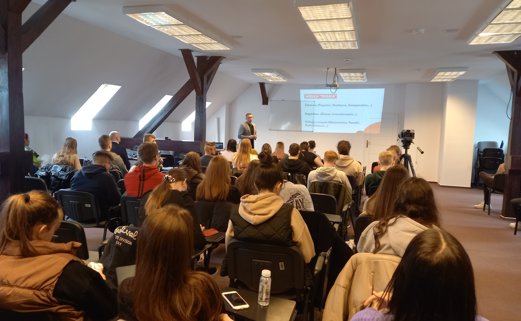 Prelegent w szarej marynarce prezentuje slajd z napisem 'WIELKA TRÓJKA' podczas wykładu dla grupy osób siedzących na krzesłach w sali z drewnianymi belkami stropowymi i oknami dachowymi.