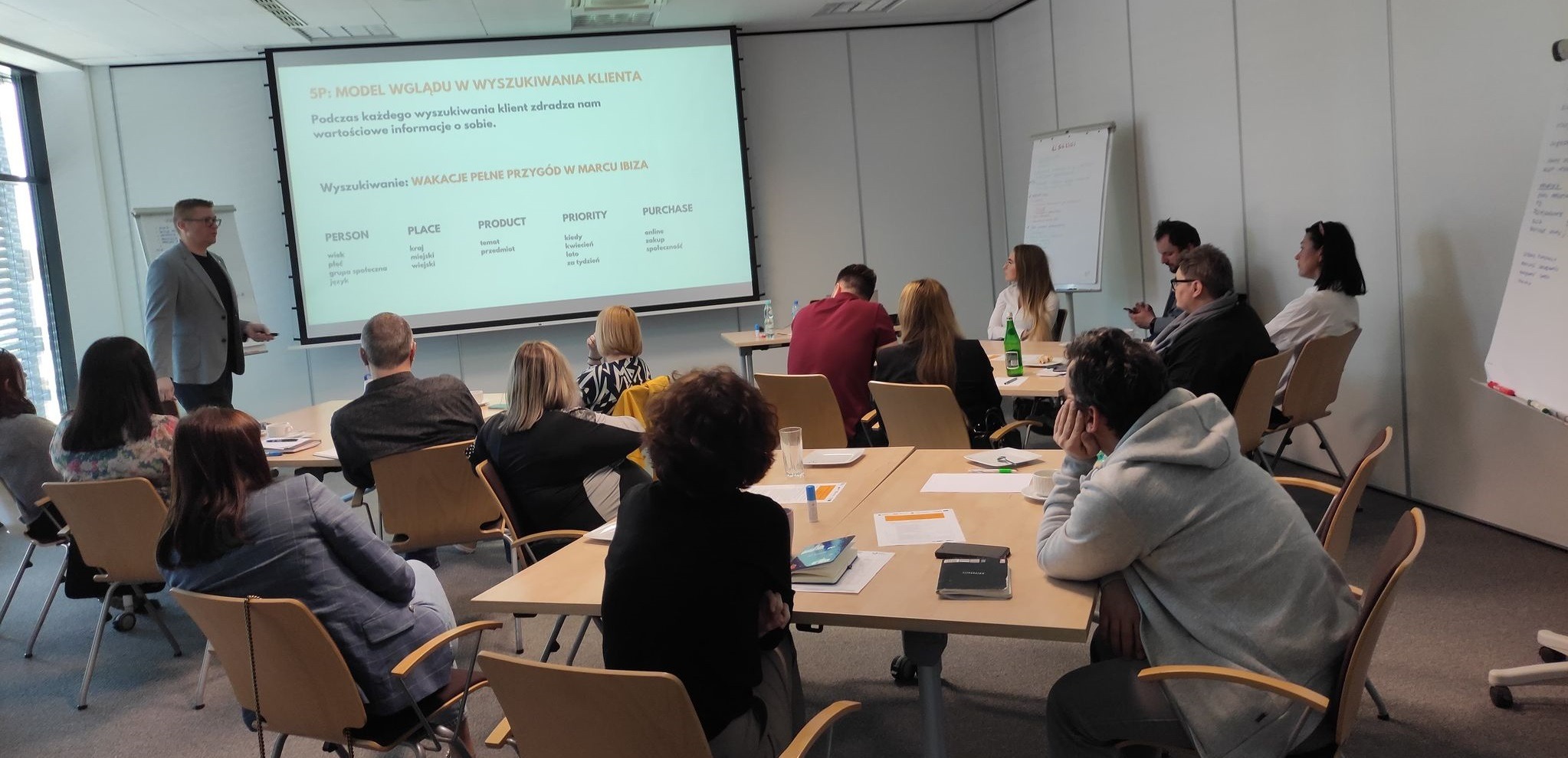 Prelegent w szarej marynarce prowadzi szkolenie z digital marketingu, prezentacja na ekranie z modelem 5P, uczestnicy siedzą przy stołach.