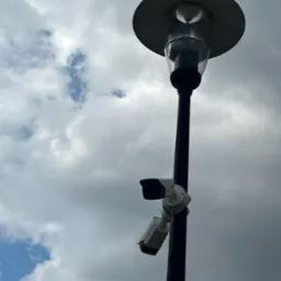 Dwie kamery monitoringu zamontowane na słupie oświetleniowym w Szczytnie, skierowane w różnych kierunkach, na tle pochmurnego nieba.