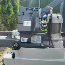 Automatyka bramy przesuwnej marki Nice R0600 z kluczem w zamku, widoczne okablowanie i silnik, na tle zieleni.