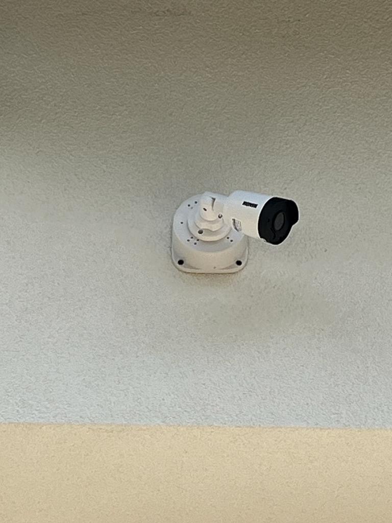 Kamera monitoringu CCTV zamontowana na jasnej, teksturowanej ścianie, widok z dołu, z widoczną podstawą montażową i czarnym obiektywem.