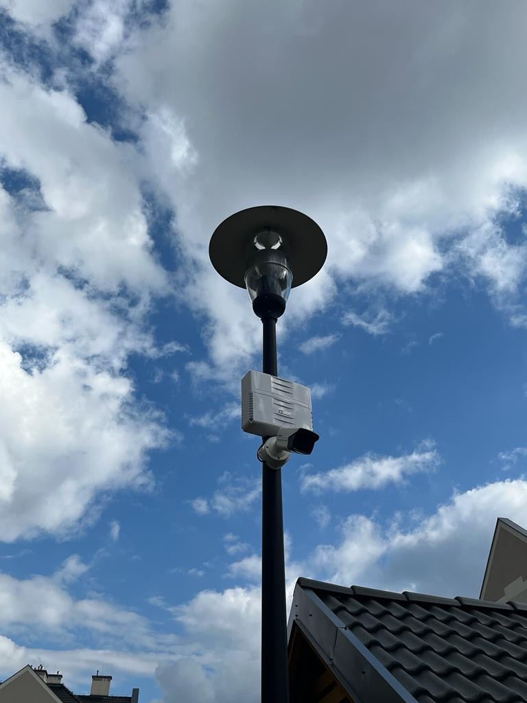 Lampa oświetleniowa z zamontowaną kamerą monitoringu i skrzynką połączeniową na tle błękitnego nieba z obłokami, widoczny fragment dachu budynku.
