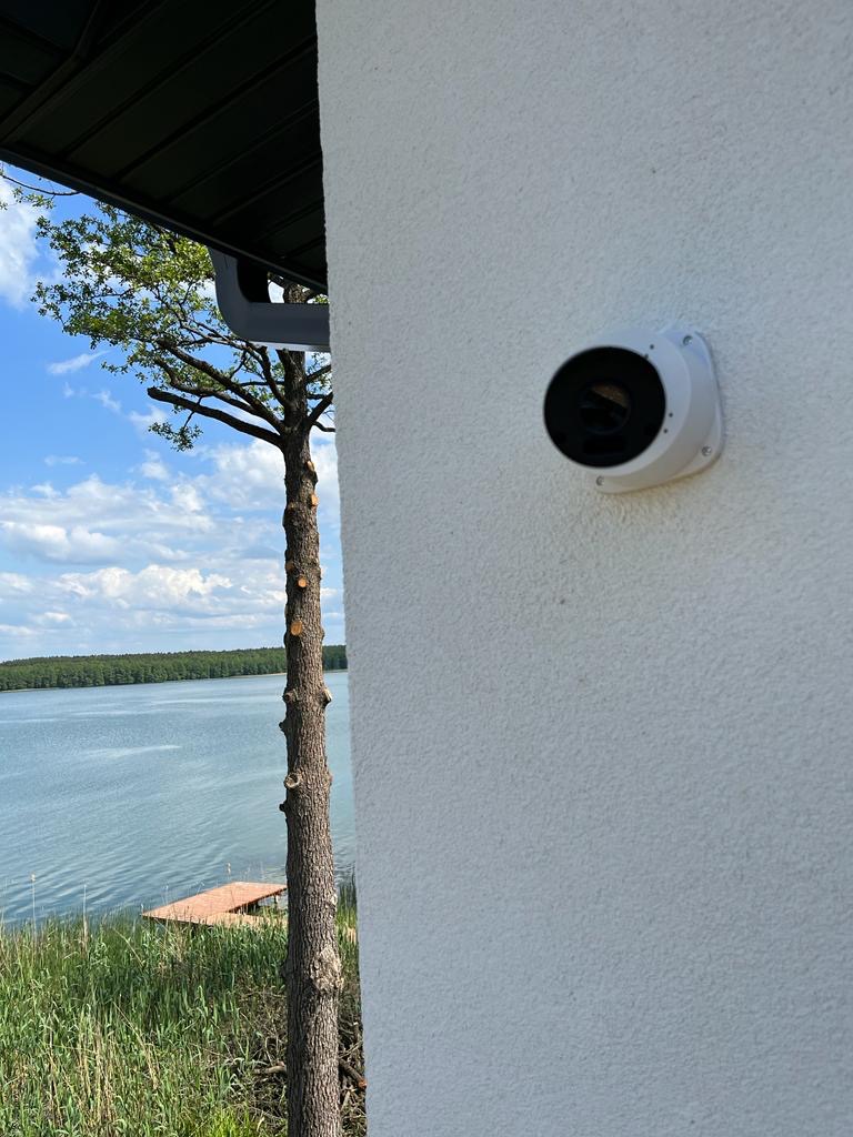 Kamera monitoringu przymocowana do białej ściany budynku z widokiem na jezioro i drzewa. Widoczny fragment dachu z rynną.