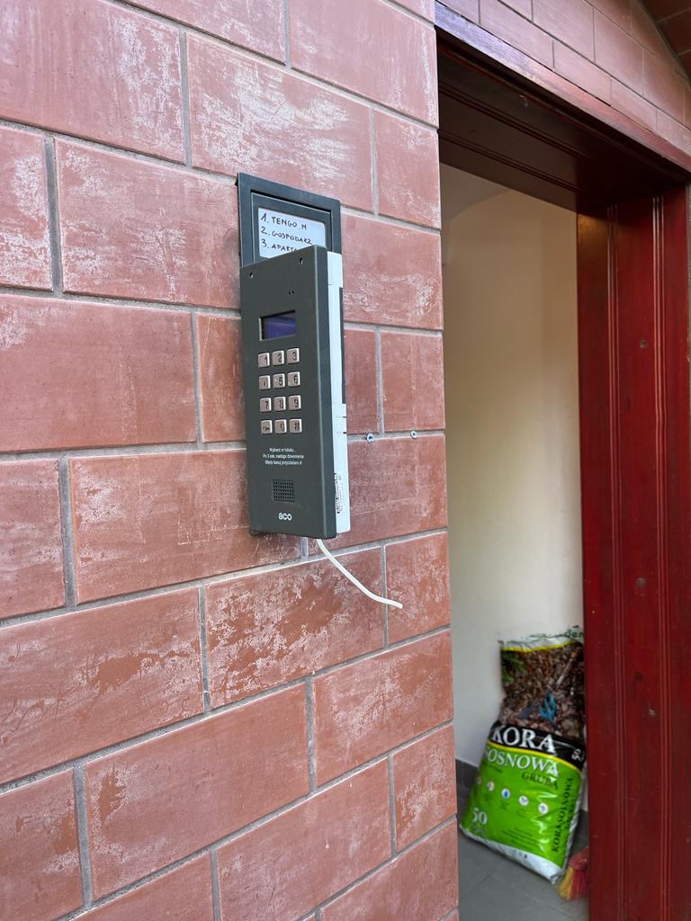 Szary domofon z klawiaturą numeryczną zamontowany na ceglanej ścianie obok tabliczki z wypisanymi odręcznie numerami mieszkań, widoczny kabel i worek kory sosnowej przy drzwiach.