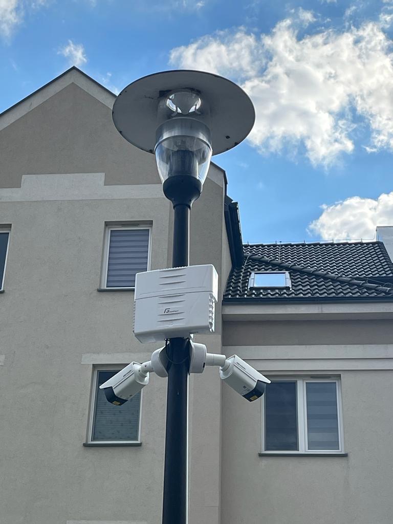 Lampa uliczna z trzema kamerami monitoringu i białą skrzynką sterującą na tle budynku mieszkalnego i błękitnego nieba z chmurami.