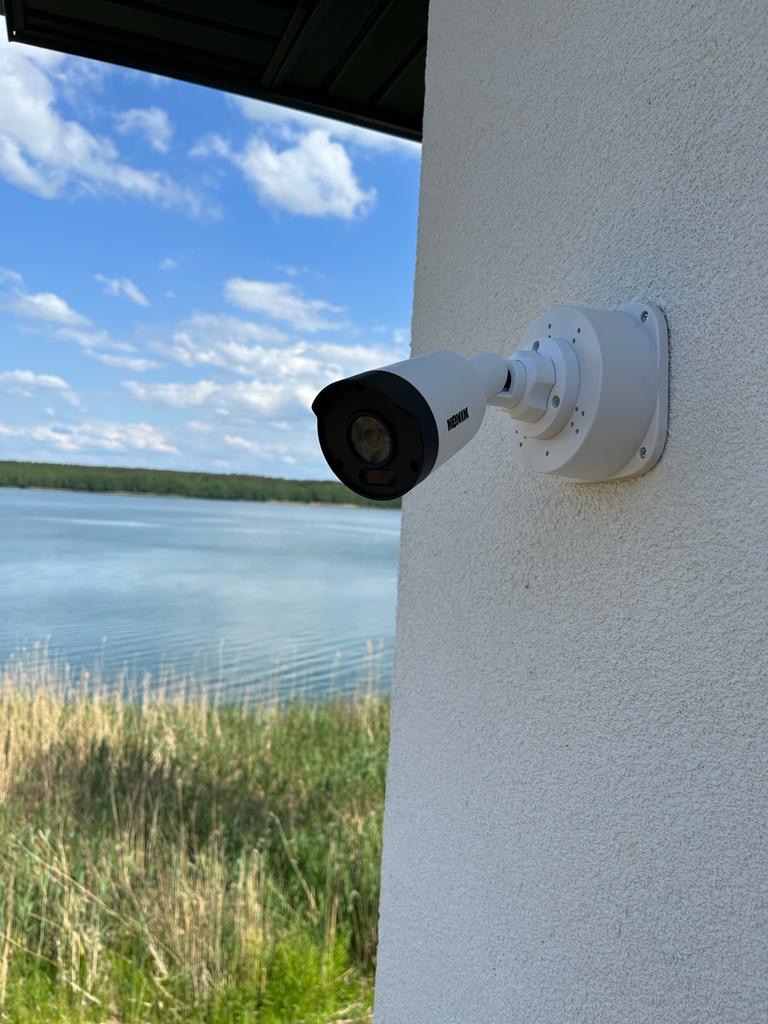 Kamera monitoringu marki HENIK zamontowana na elewacji budynku z widokiem na jezioro i trzciny w tle.