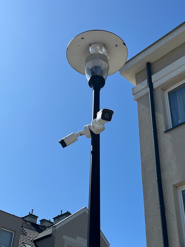 Dwie kamery monitoringu zamontowane na słupie oświetleniowym, skierowane w różnych kierunkach, na tle błękitnego nieba i fragmentów budynków mieszkalnych.