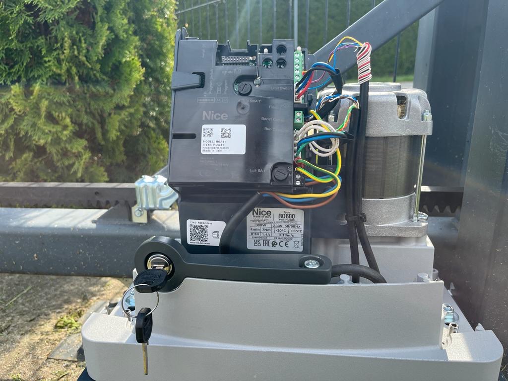 Automatyka bramy przesuwnej marki Nice R0600 z kluczem w zamku, widoczne okablowanie i silnik, na tle zieleni.