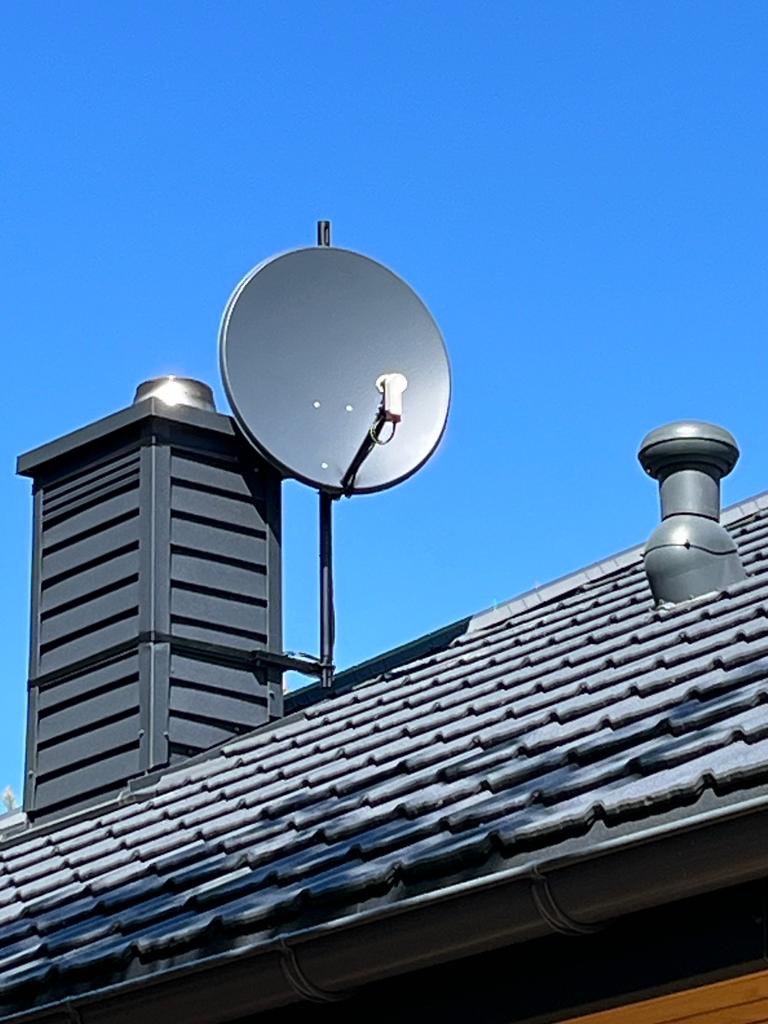 Antena satelitarna zamontowana na dachu domu, widoczna obok komina i wentylacji, na tle błękitnego nieba.