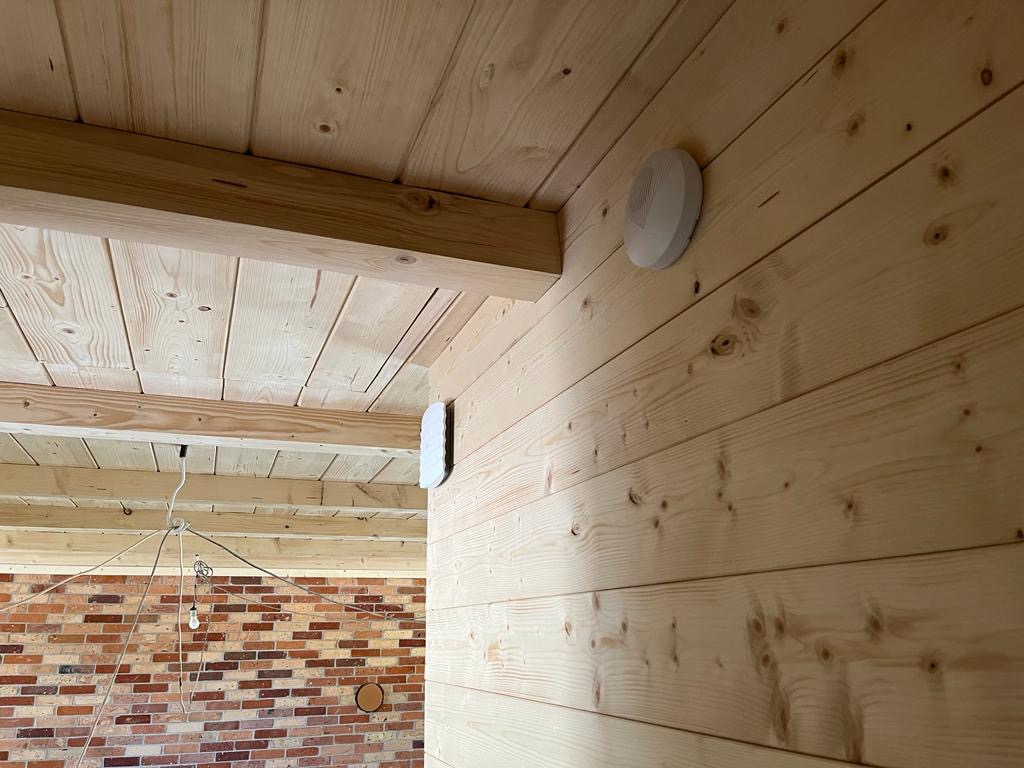 Wnętrze wykończone jasnym drewnem boazeryjnym z widocznymi dwoma okrągłymi czujnikami na ścianie, z fragmentem muru z czerwonej cegły w tle.