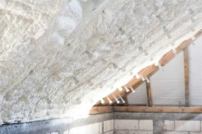 Szczegółowe ujęcie izolacji poddasza pianką PUR z widocznymi plastikowymi kołkami mocującymi do drewnianej konstrukcji dachu, w tle fragment ściany z bloczków.