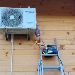 AK Serwis - Biała jednostka zewnętrzna klimatyzatora Whirlpool Inverter Plus zamontowana na drewnianej ścianie domu, podłączona do pompy próżniowej stojącej na aluminiowej drabinie.