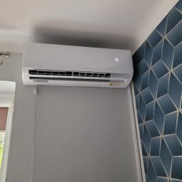 AK Serwis - Biała jednostka klimatyzacji ściennej z cyfrowym wyświetlaczem temperatury '16', zamontowana w pokoju z szarą ścianą i granatową tapetą w geometryczne wzory; widoczne okno z zasłoną i zielenią za...