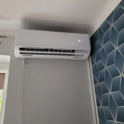 Biała jednostka klimatyzacji ściennej z cyfrowym wyświetlaczem temperatury '16', zamontowana w pokoju z szarą ścianą i granatową tapetą w geometryczne wzory; widoczne okno z zasłoną i zielenią za...