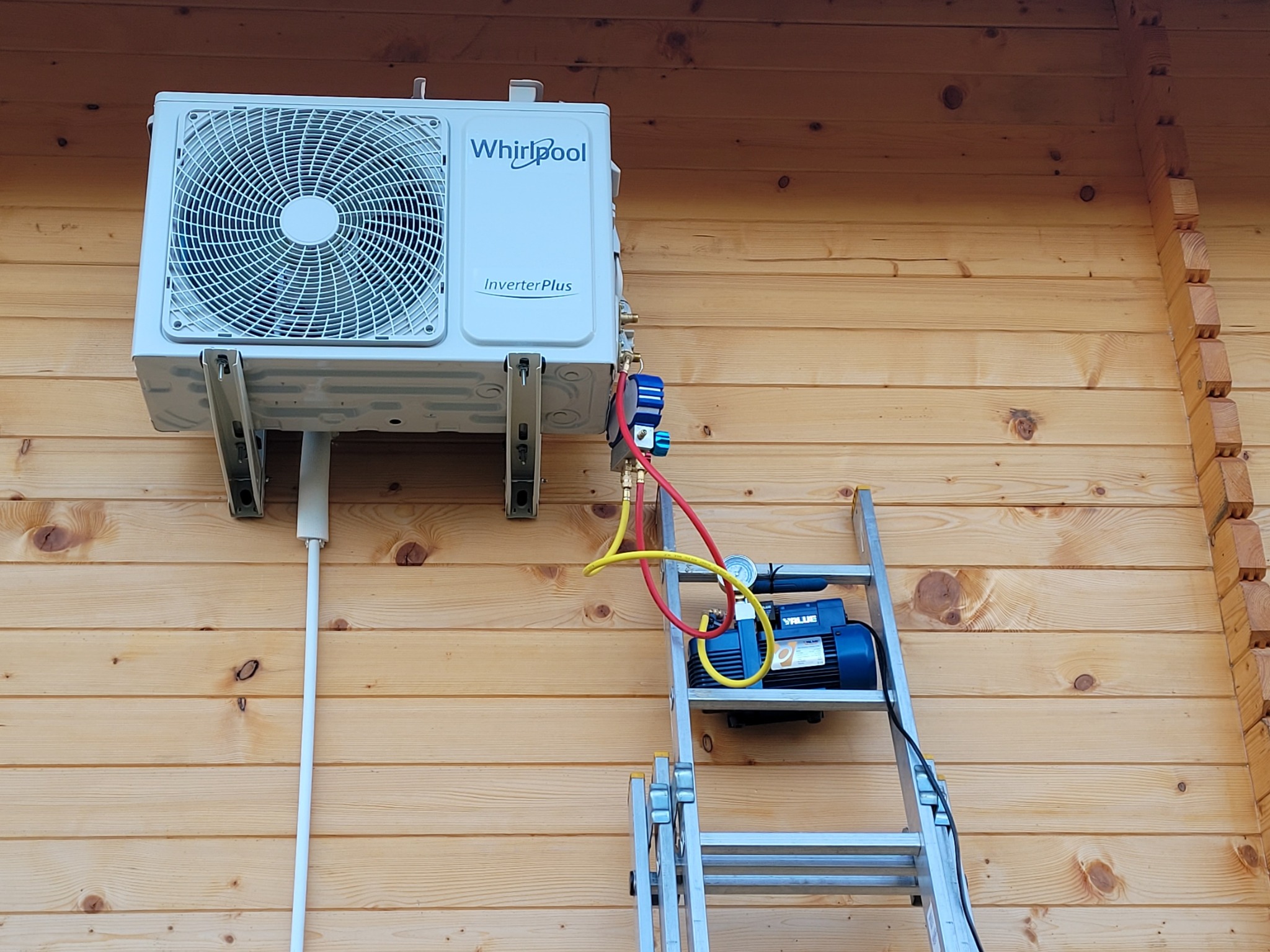 Biała jednostka zewnętrzna klimatyzatora Whirlpool Inverter Plus zamontowana na drewnianej ścianie domu, podłączona do pompy próżniowej stojącej na aluminiowej drabinie.