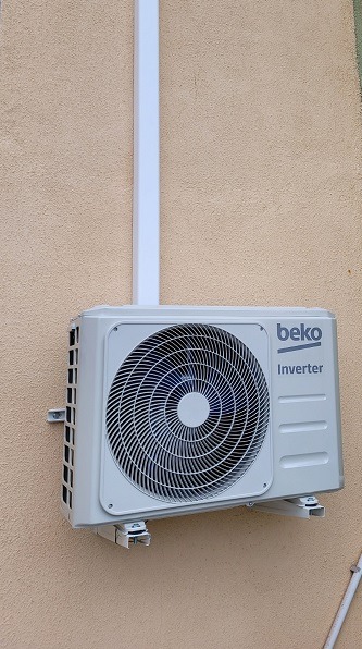 Zewnętrzna jednostka klimatyzacji Beko Inverter zamontowana na beżowej, teksturowanej ścianie, z białą rurą prowadzącą w górę.