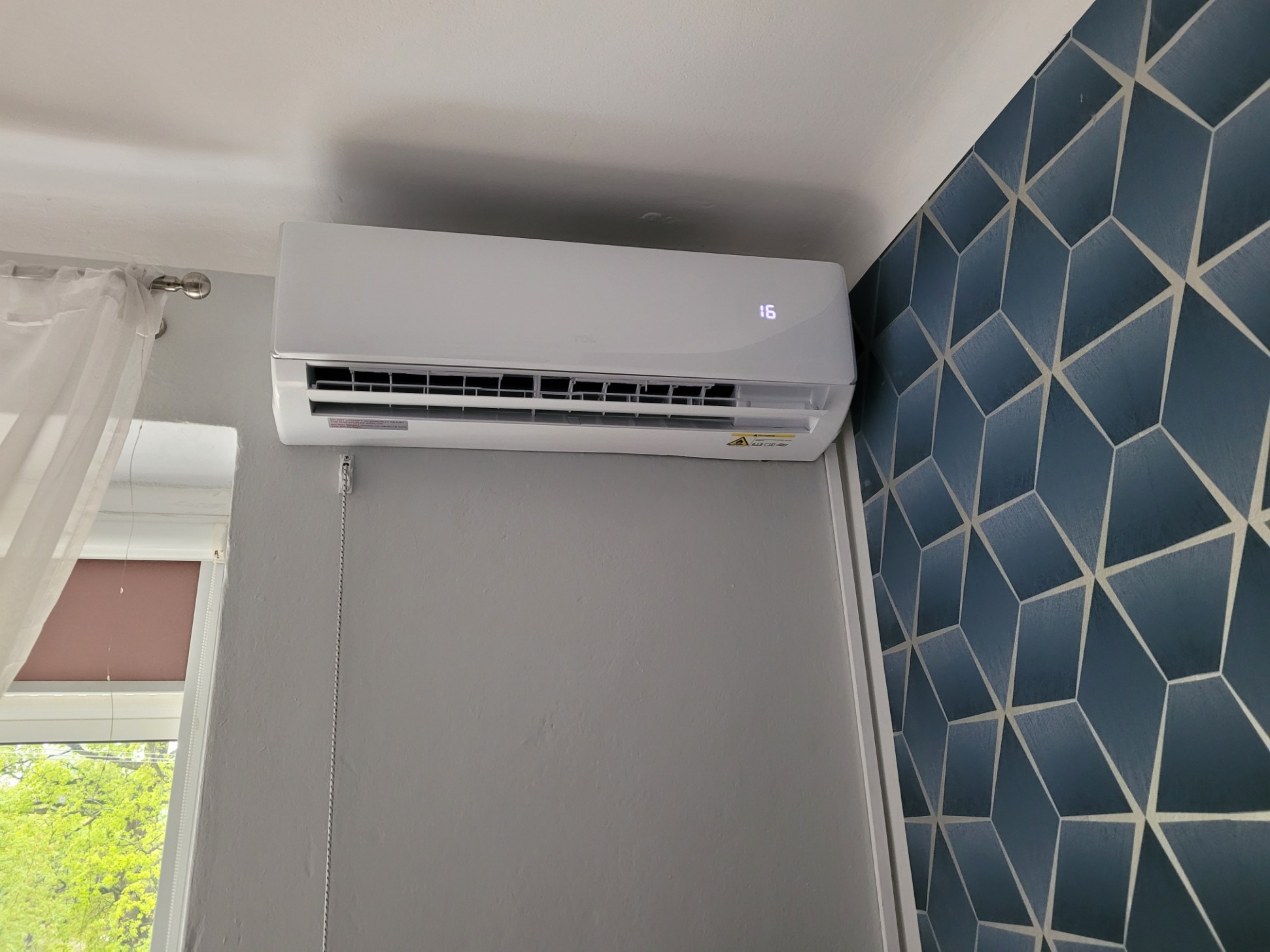Biała jednostka klimatyzacji ściennej z cyfrowym wyświetlaczem temperatury '16', zamontowana w pokoju z szarą ścianą i granatową tapetą w geometryczne wzory; widoczne okno z zasłoną i zielenią za...