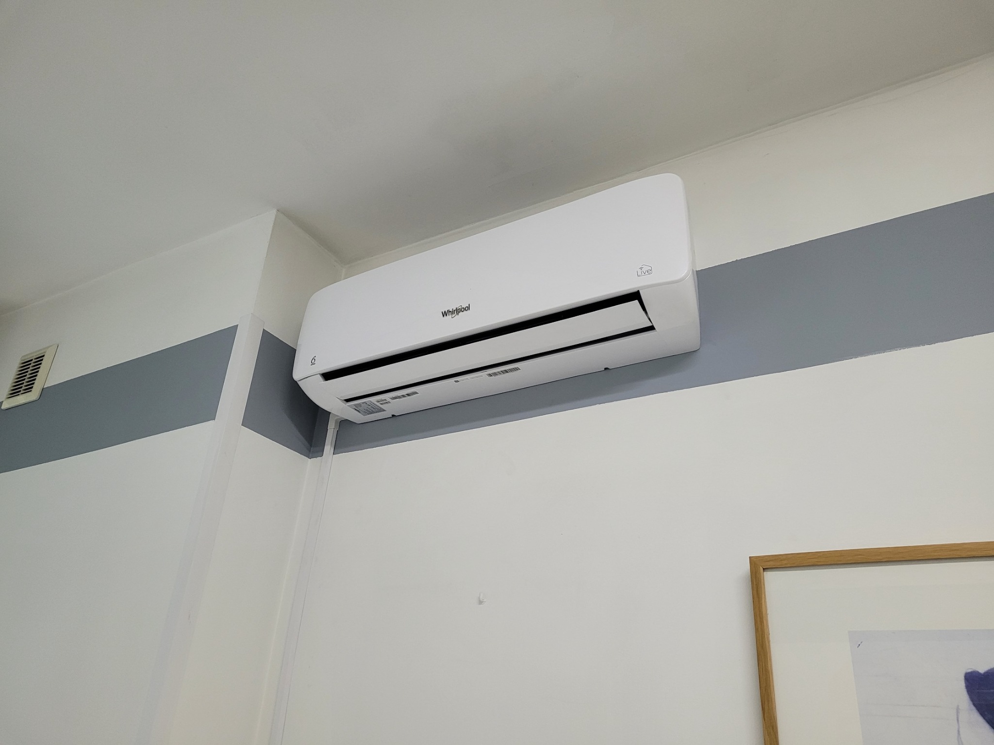 Biały klimatyzator Whirlpool zamontowany na ścianie z szarym pasem, widoczny fragment obrazu w ramie.
