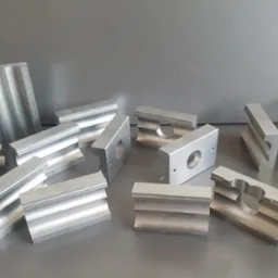 Surowe, aluminiowe elementy obrobione w Brz&oacute;ze, z widocznymi śladami frezowania i otworami, ułożone na szarym tle.