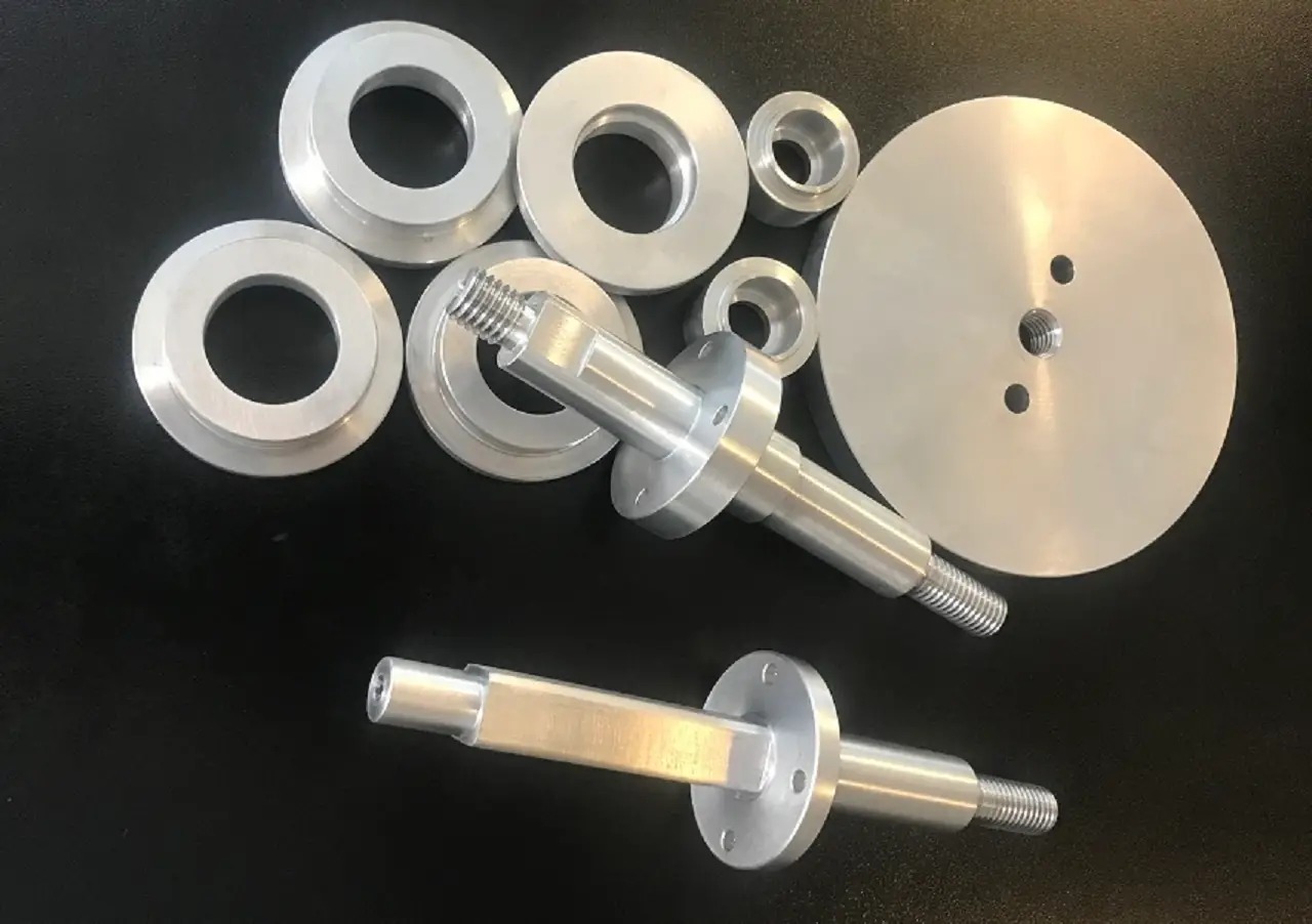 Precyzyjnie obrobione, srebrne elementy metalowe, w tym gwintowane trzpienie i pierścienie, ułożone na czarnym tle, ukazujące detale obróbki i gwintów.