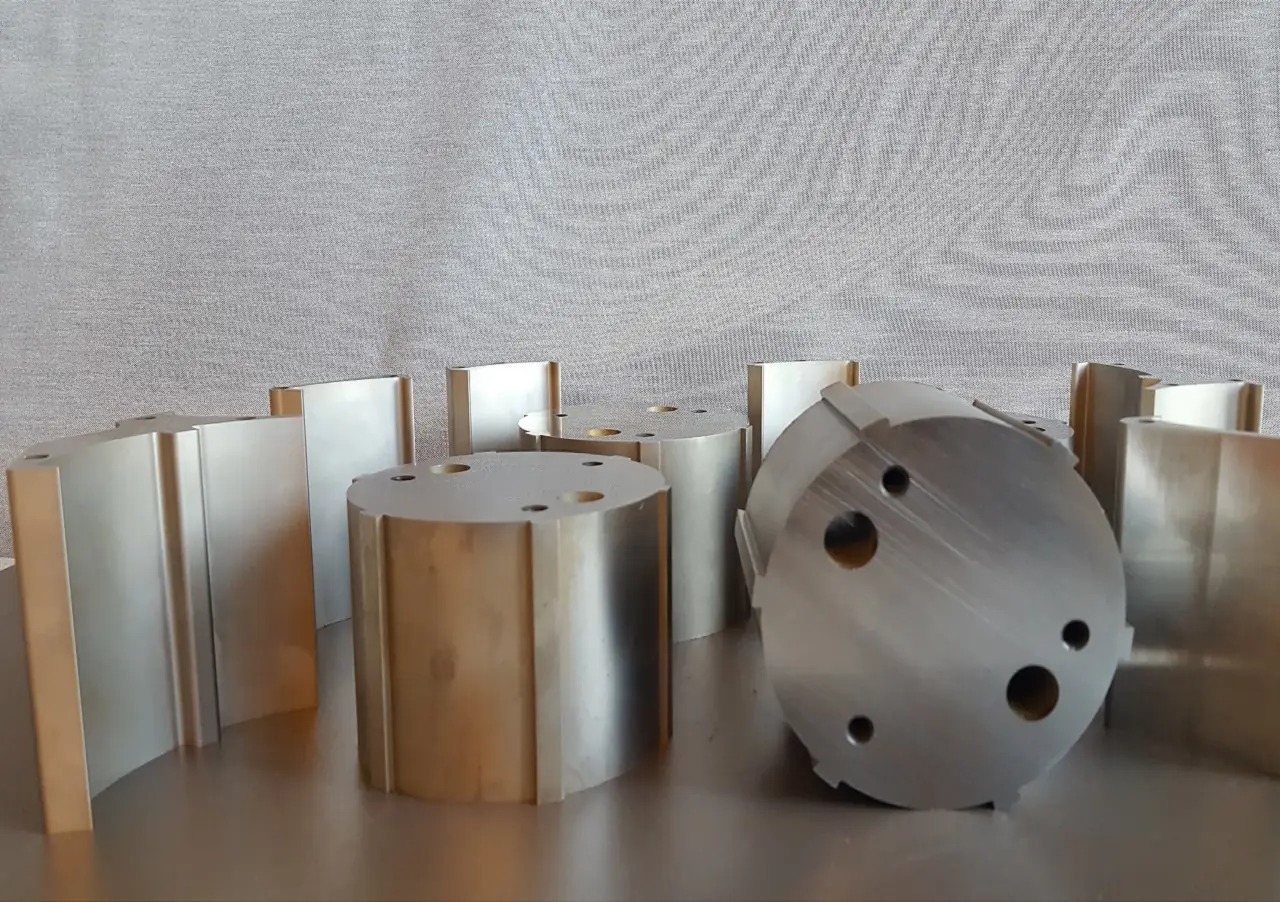 Precyzyjnie obrobione metalowe elementy cylindryczne i prostopadłościenne z otworami, ustawione na metalowej powierzchni na tle szarej tkaniny.