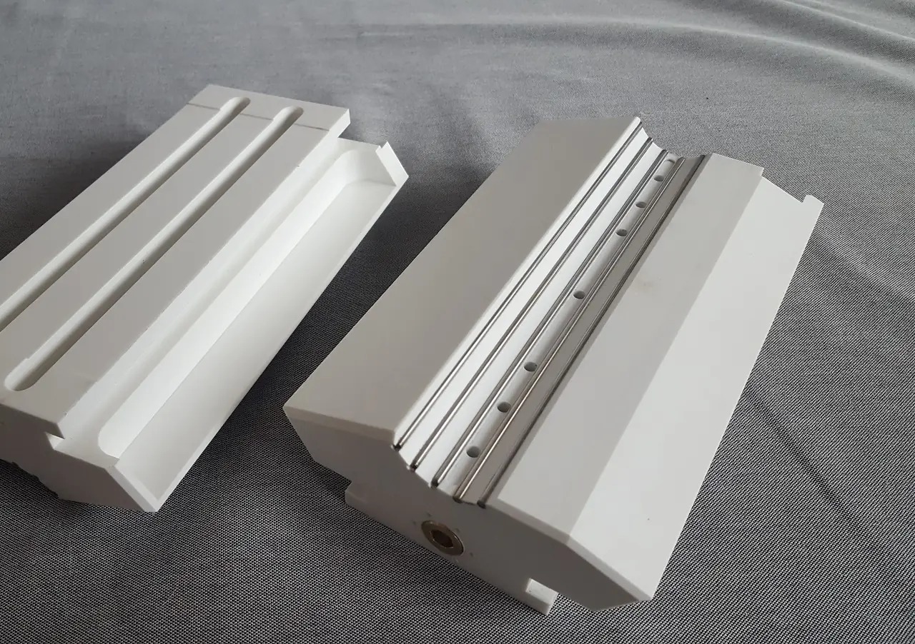Dwa białe, precyzyjnie wykonane elementy z metalowymi prętami i otworami, prawdopodobnie fragmenty konstrukcji metalowej, leżące na szarej powierzchni.