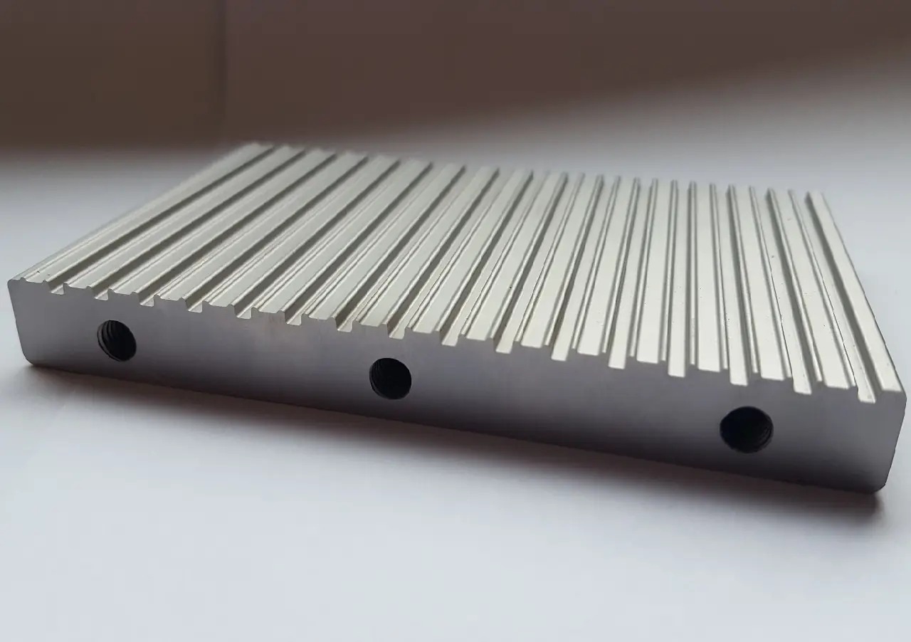 Metalowy element z równoległymi żebrami i trzema otworami na boku, obrobiony maszynowo, leżący na białym tle.