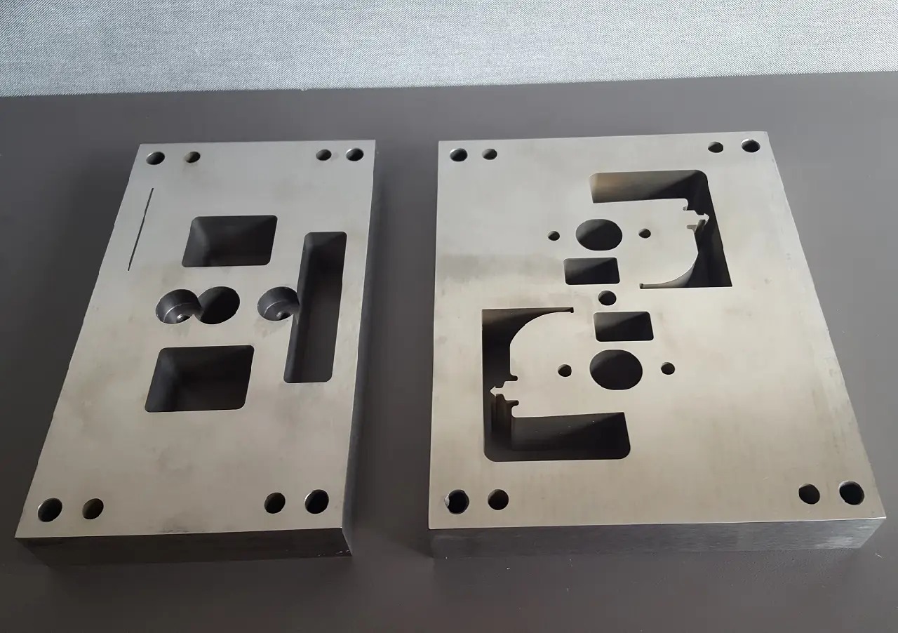 Dwie metalowe płyty z precyzyjnie wyciętymi otworami o różnych kształtach, leżące na szarej powierzchni. Widoczne otwory okrągłe, kwadratowe oraz o nieregularnych kształtach. Płyty o gładkiej...