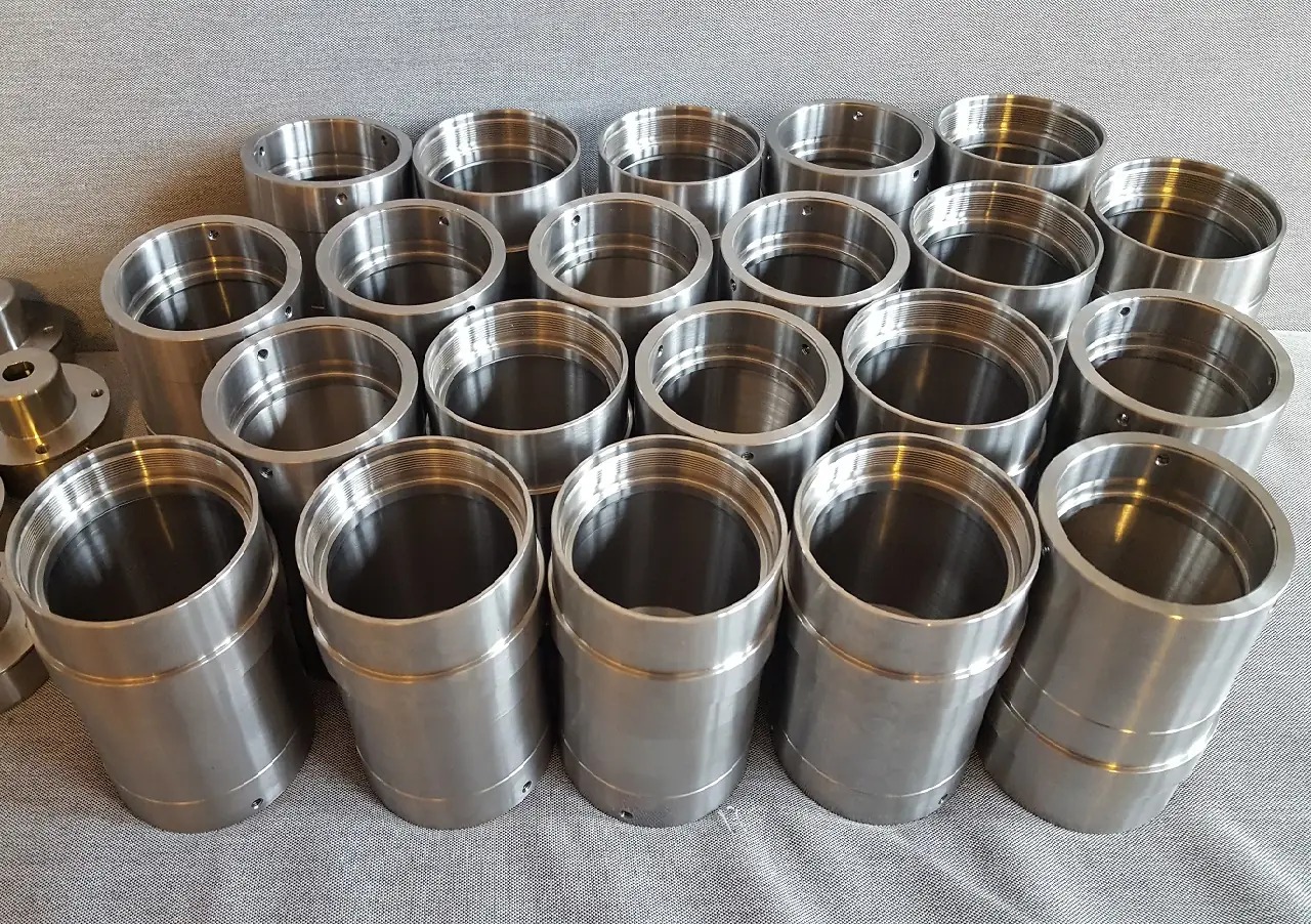 Partia cylindrycznych, metalowych elementów z gwintem wewnętrznym i otworami, ułożonych na szarym tle.