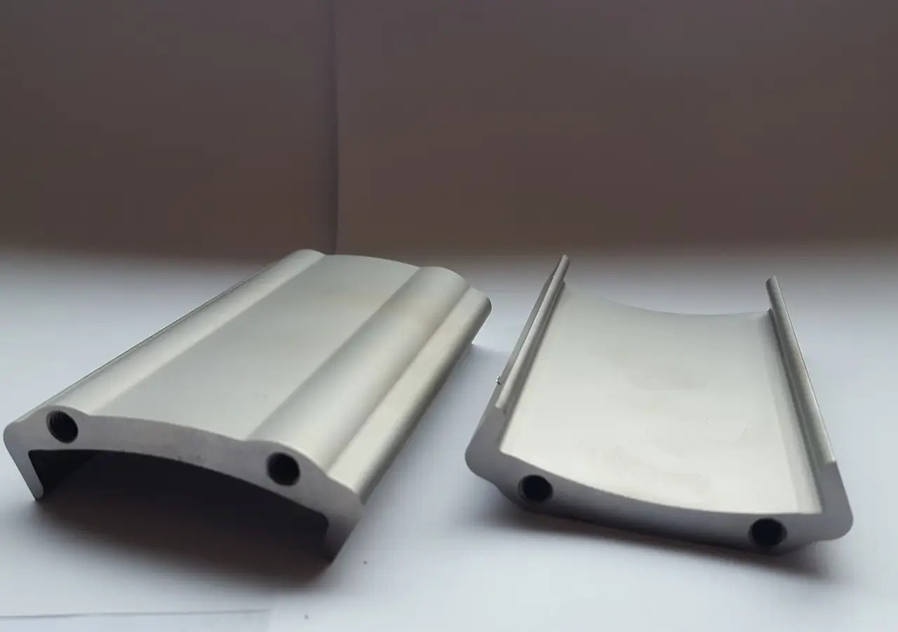 Dwa aluminiowe profile o przekroju półksiężyca z otworami, leżące na białym tle, prezentacja detali wykończeniowych.