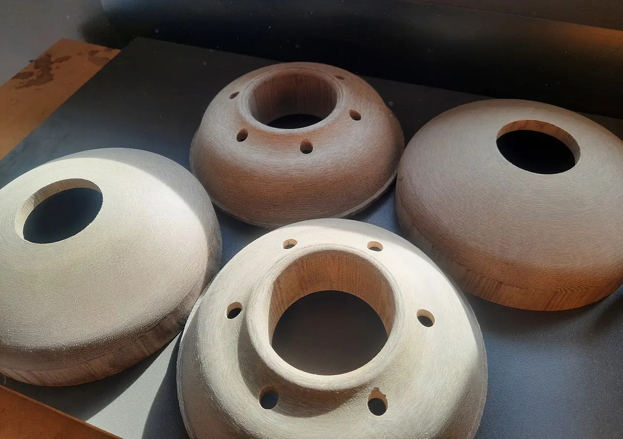 Cztery drewniane, okrągłe elementy z otworami, prawdopodobnie półprodukty do lamp lub innych elementów dekoracyjnych, leżące na ciemnym blacie, oświetlone z lewej strony.